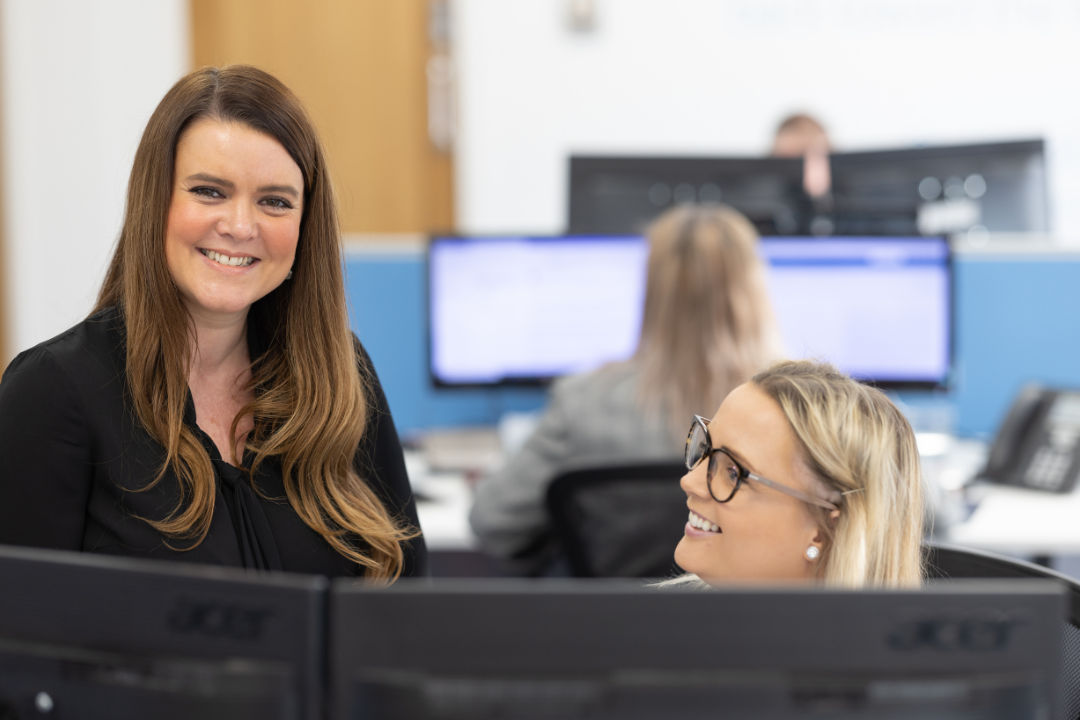 Two women sat behind computers smiling